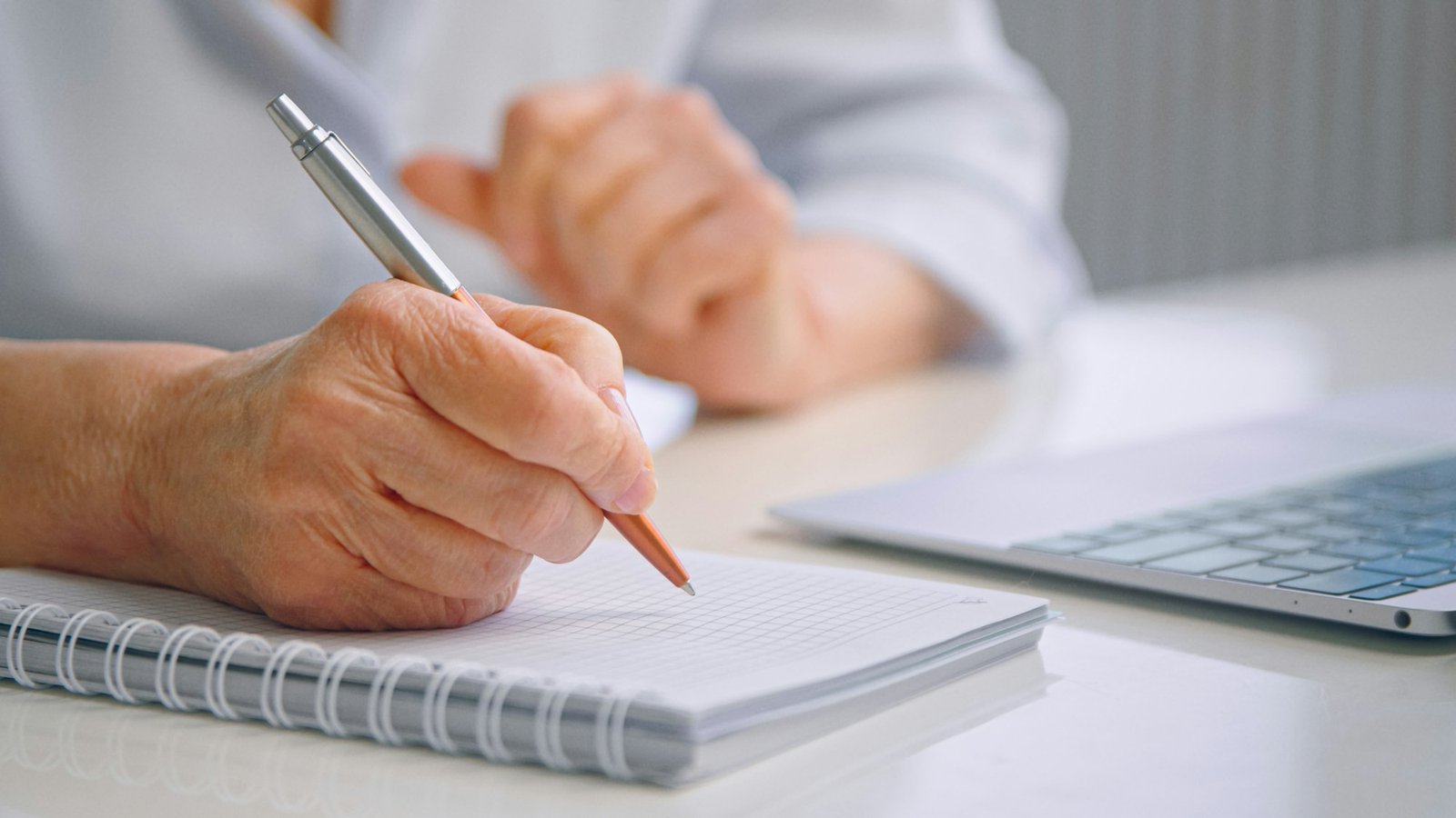 Senior lady teacher with wrinkly hands holds coloured pen over white page of paper notebook